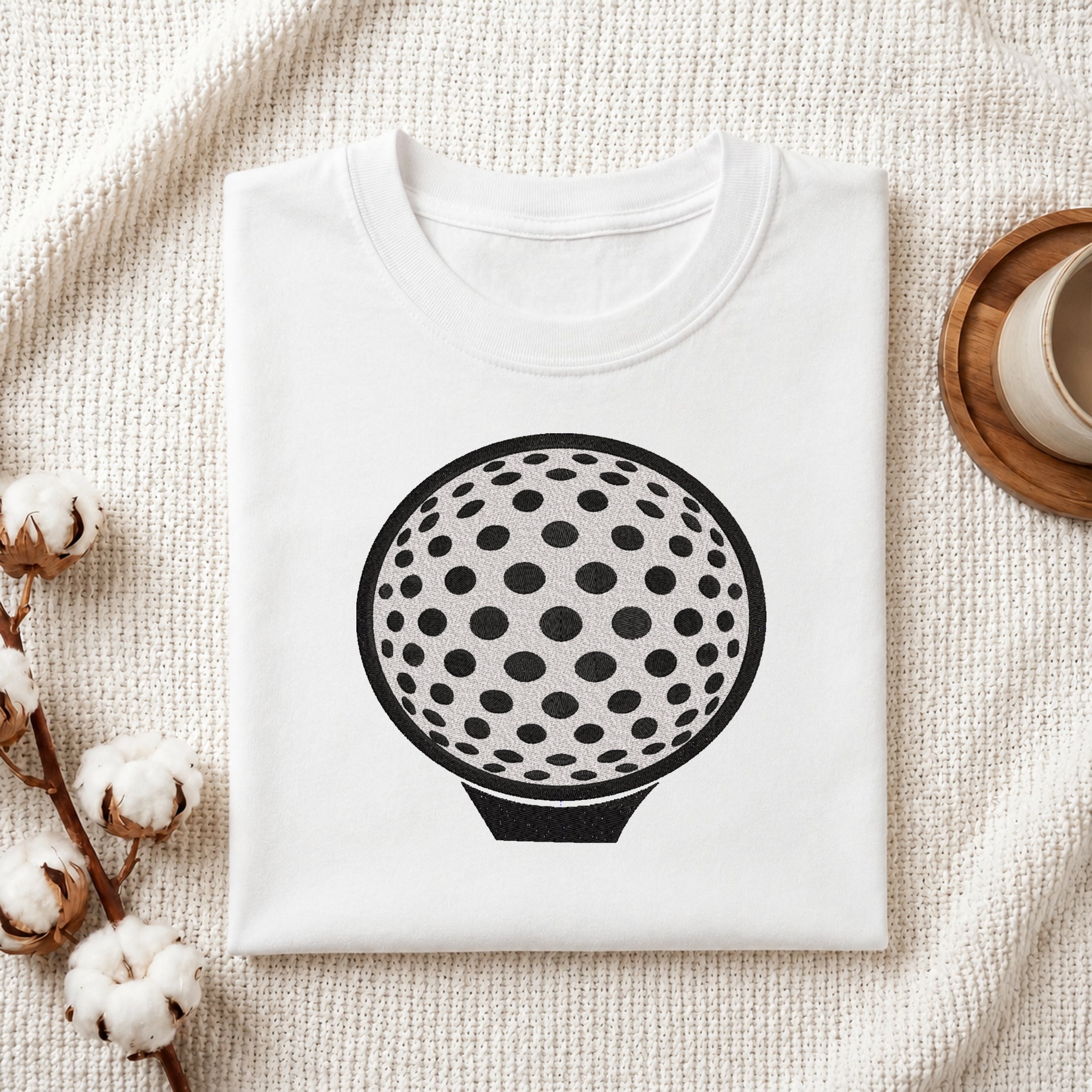A folded white crewneck t-shirt featuring a black graphic of a golf ball with black spots, styled on a textured cream fabric with cotton stems and a wooden cup in the background.
