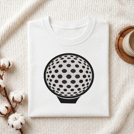 A folded white crewneck t-shirt featuring a black graphic of a golf ball with black spots, styled on a textured cream fabric with cotton stems and a wooden cup in the background.