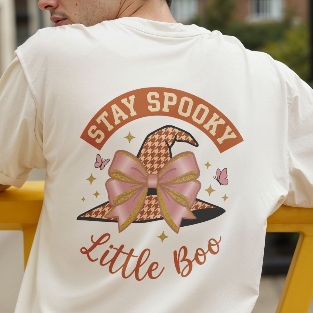 A person wearing a cream-colored t-shirt with a large 'Stay Spooky Little Boo' graphic on the back, featuring a witch hat with a pink bow and houndstooth pattern.