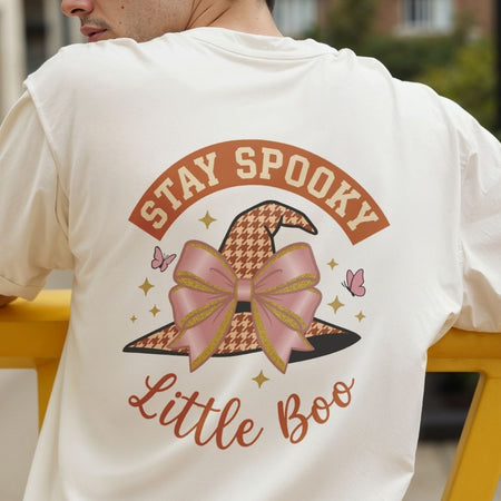 A person wearing a cream-colored t-shirt with a large 'Stay Spooky Little Boo' graphic on the back, featuring a witch hat with a pink bow and houndstooth pattern.