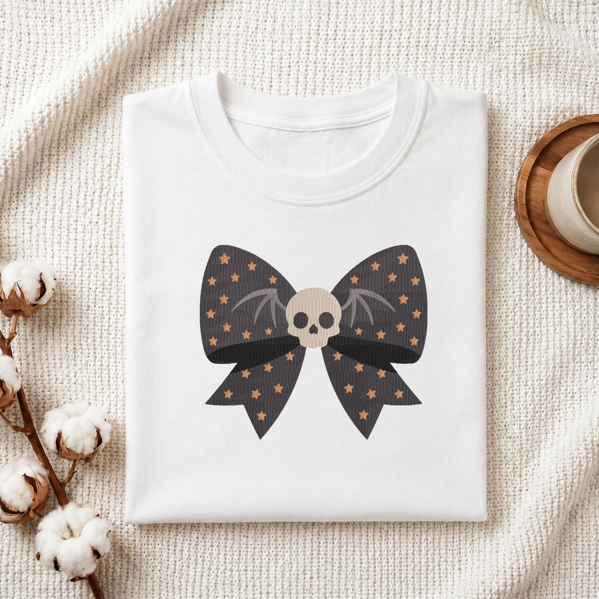 A neatly folded white t-shirt featuring the embroidered starry bow and skull design, displayed on a textured white knit background with cotton stems and a wooden cup.