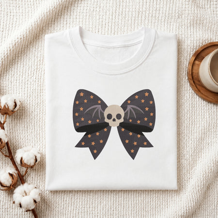 A neatly folded white t-shirt featuring the embroidered starry bow and skull design, displayed on a textured white knit background with cotton stems and a wooden cup.