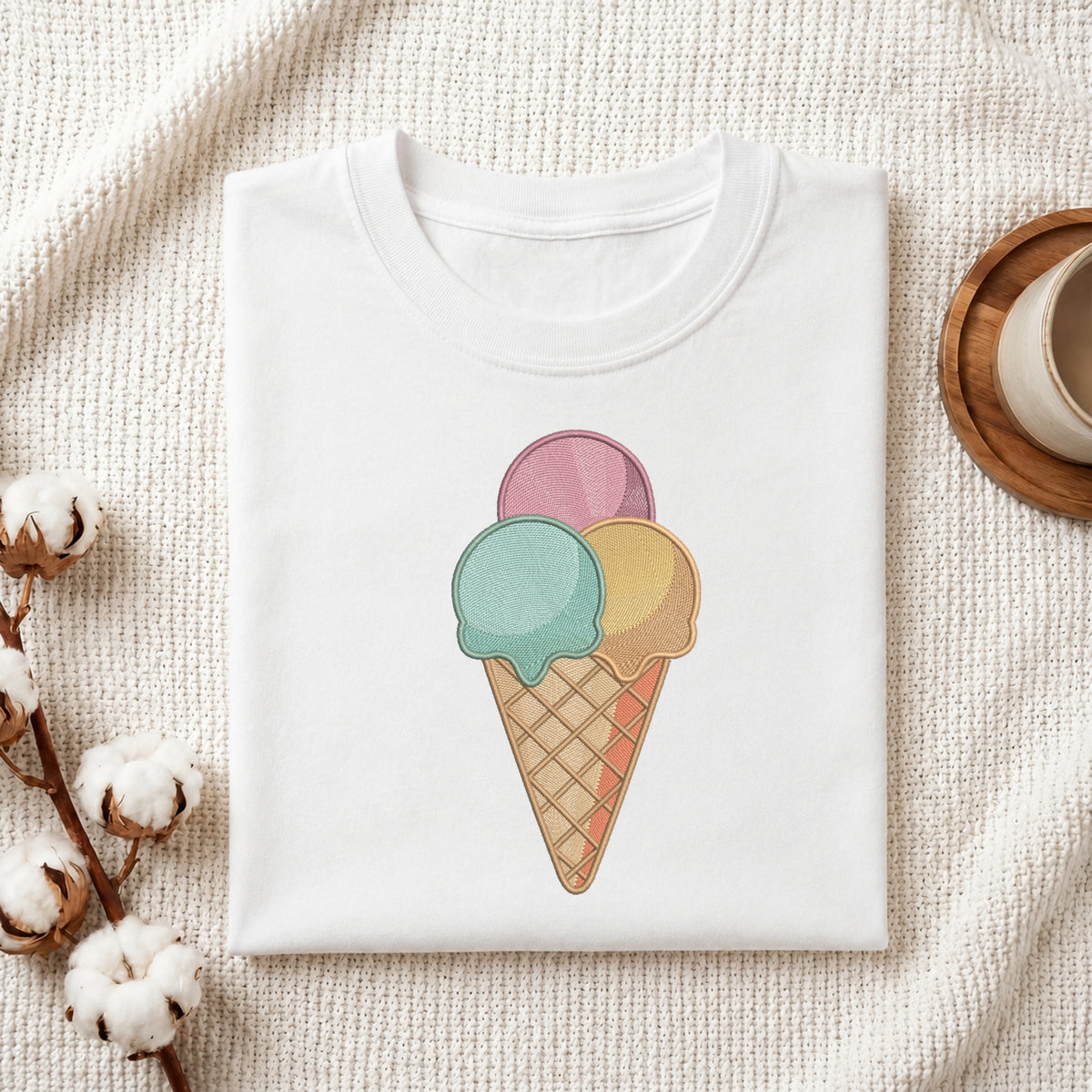 A folded white T-shirt with a pastel ice cream cone graphic on the front, styled on a textured cream fabric with cotton stems and a small cup at the side.