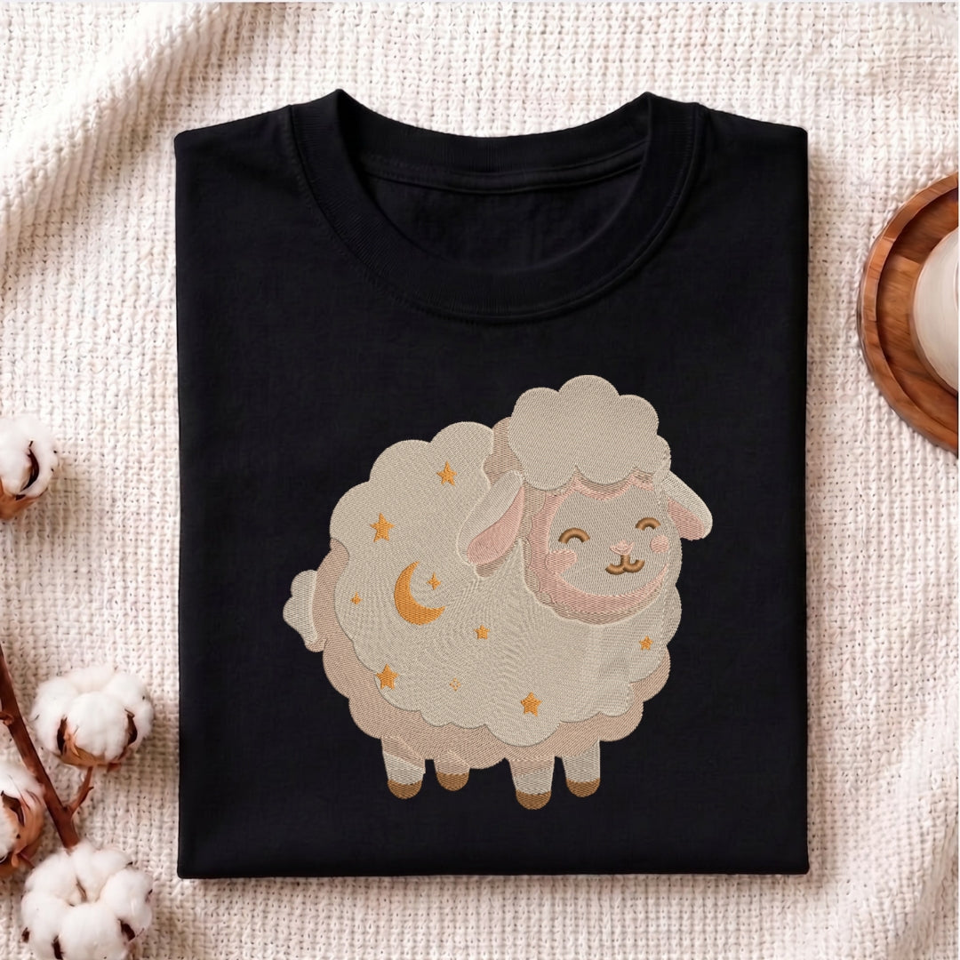 A folded black crewneck t-shirt featuring a cute cartoon sheep graphic with embroidered stars and a crescent moon, displayed on a textured beige fabric background with cotton bolls and a wooden coaster.