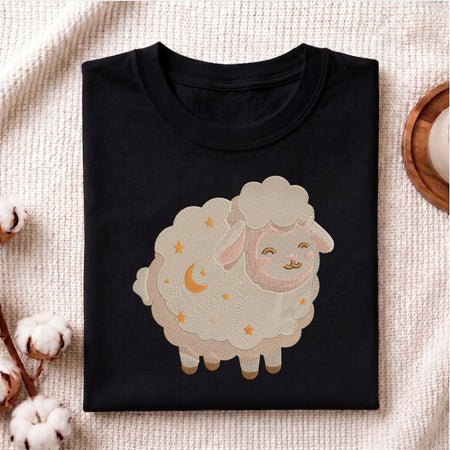 A folded black crewneck t-shirt featuring a cute cartoon sheep graphic with embroidered stars and a crescent moon, displayed on a textured beige fabric background with cotton bolls and a wooden coaster.
