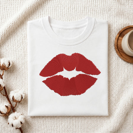 A white folded t-shirt featuring a detailed, embroidered red lips design, presented on a textured white fabric with cotton sprigs and a wooden coaster.