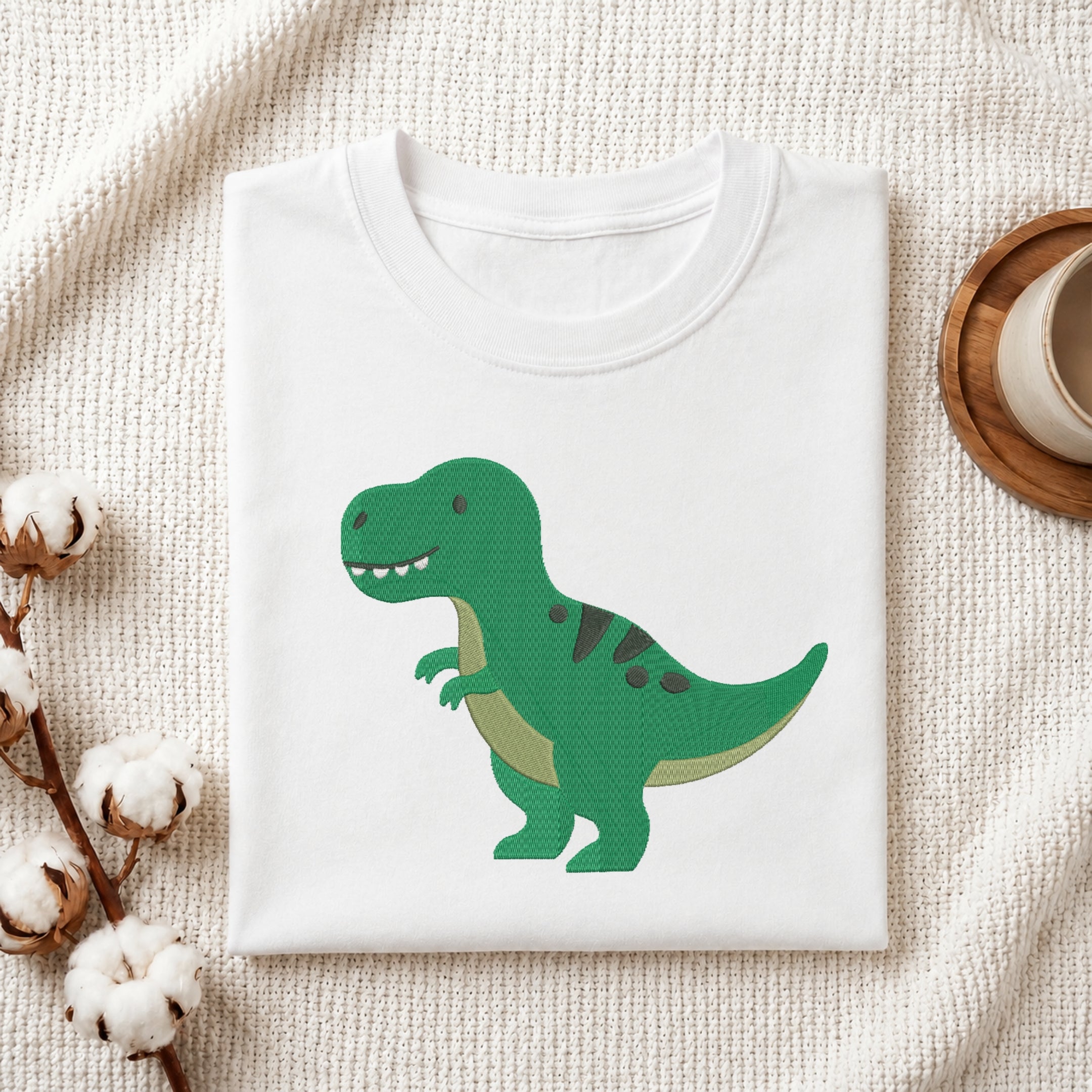 A folded white crewneck t-shirt featuring a cute green cartoon T-Rex dinosaur graphic, styled on a textured cream knit blanket with cotton bolls and a wooden cup in the background.