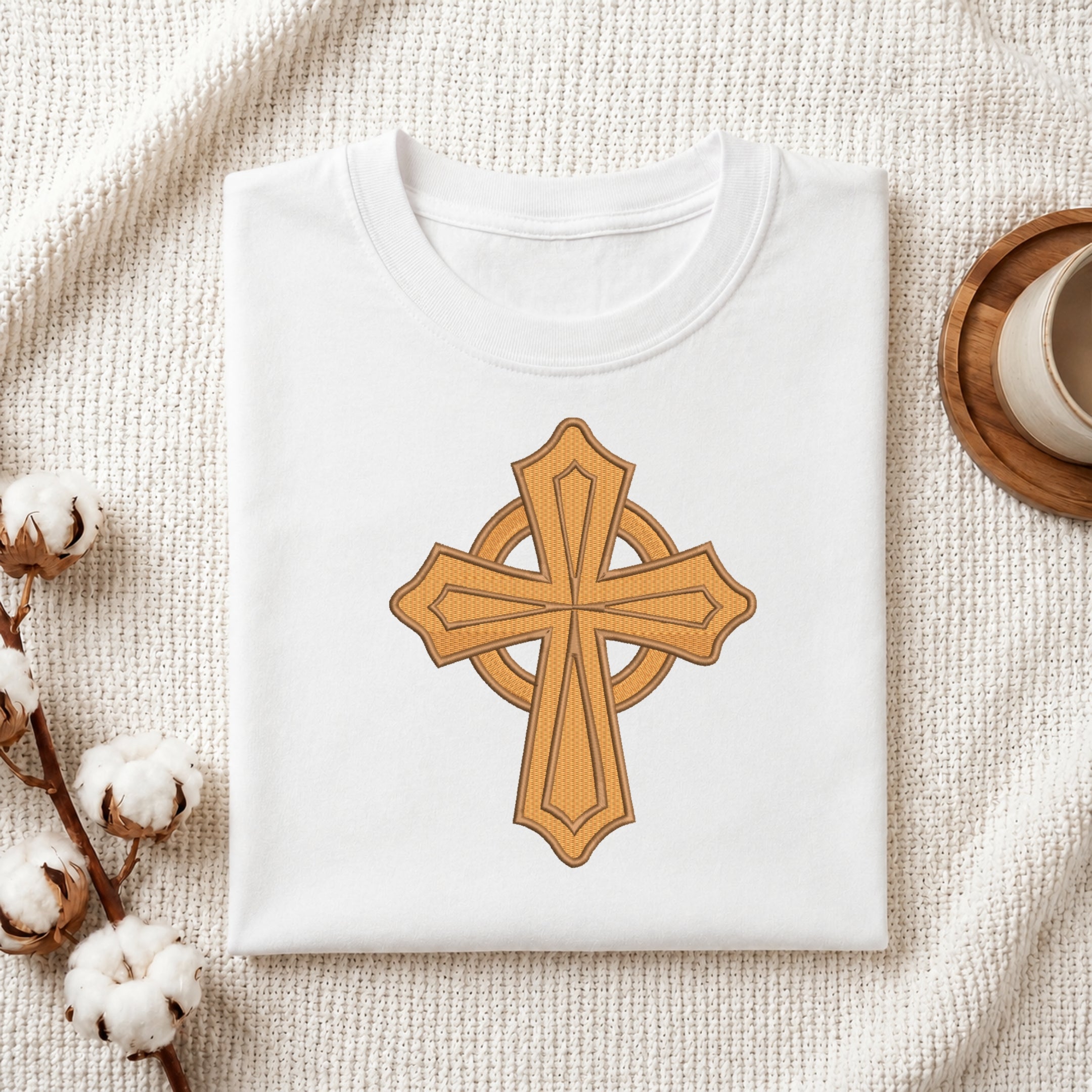 A folded white t-shirt with a golden-brown Celtic cross graphic printed on the front, styled on a textured cream knit blanket with cotton bolls and a wooden cup in the background.