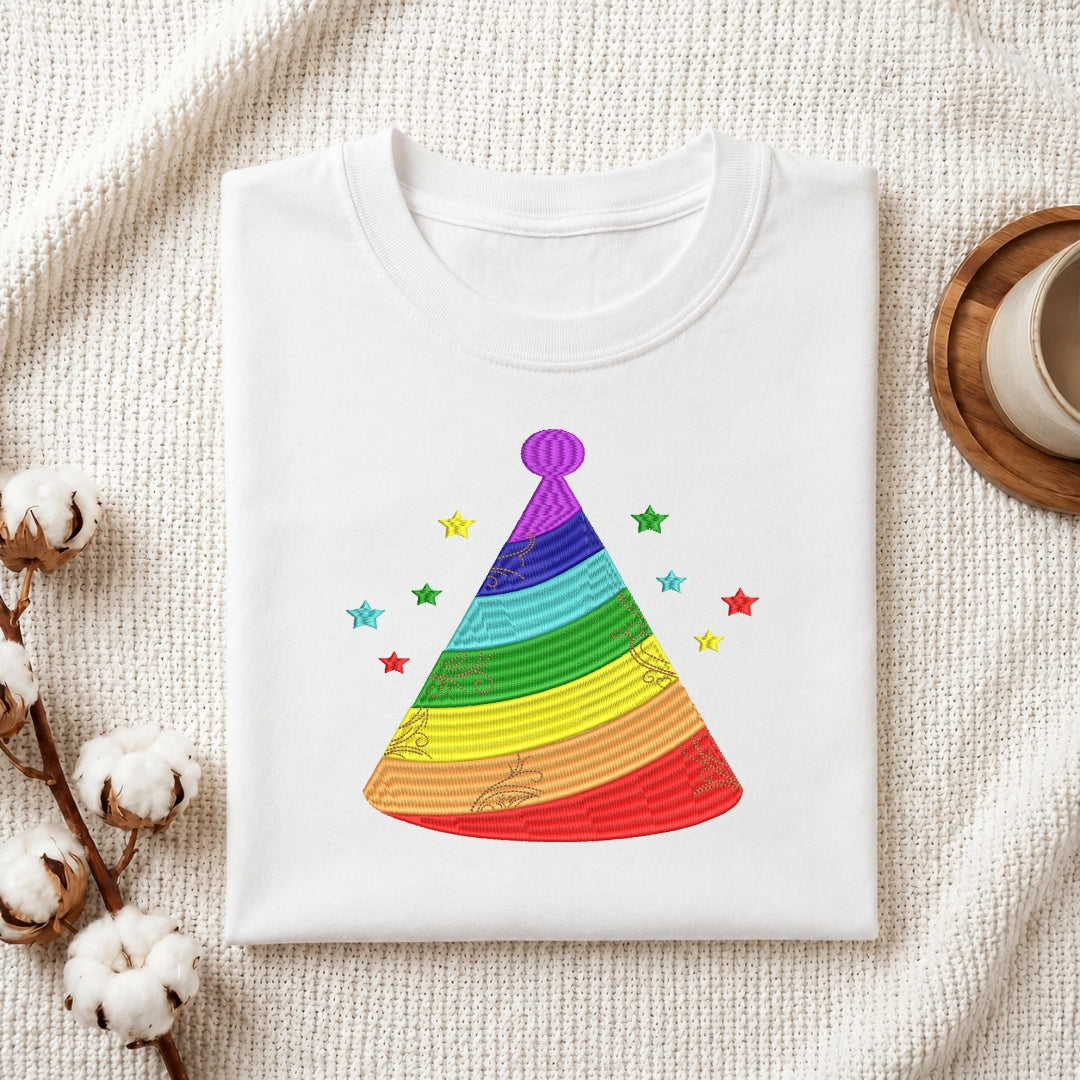 A folded white t-shirt with a colorful rainbow party hat graphic and small stars, displayed on a textured cream fabric with cotton bolls and a cup in the background.