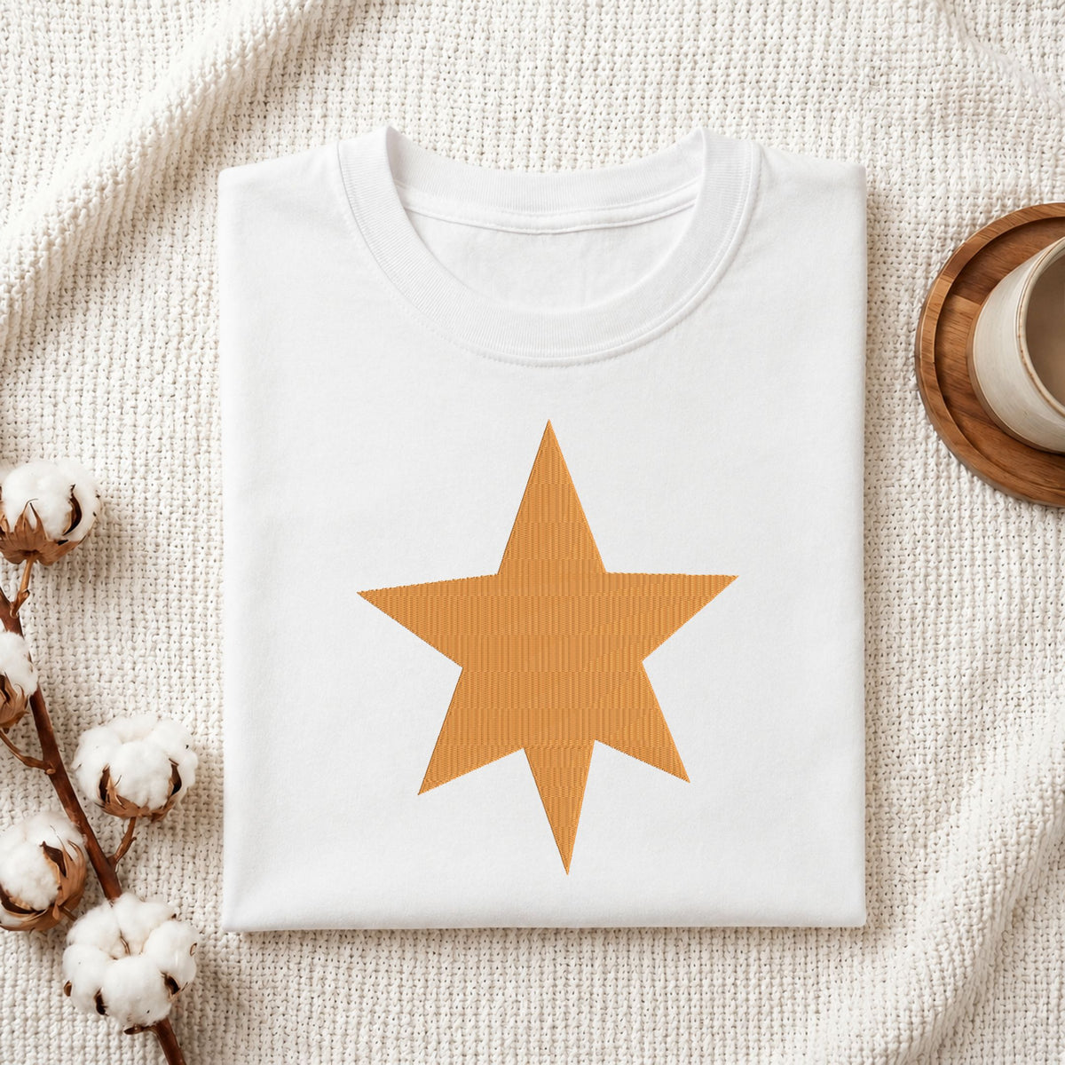 Folded white cotton t-shirt mockup featuring a seven-pointed gold star machine embroidery design, styled with cotton stems and a wooden cup.

