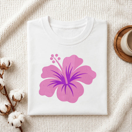 A folded white crewneck t-shirt featuring a large pink hibiscus flower graphic with a purple center, styled on a textured cream knit blanket with cotton bolls and a wooden cup in the background.