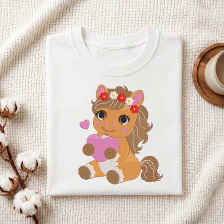 A folded white crewneck t-shirt featuring a cute cartoon illustration of a brown pony with a pink heart and floral headband, styled on a textured cream fabric with cotton bolls and a wooden cup.