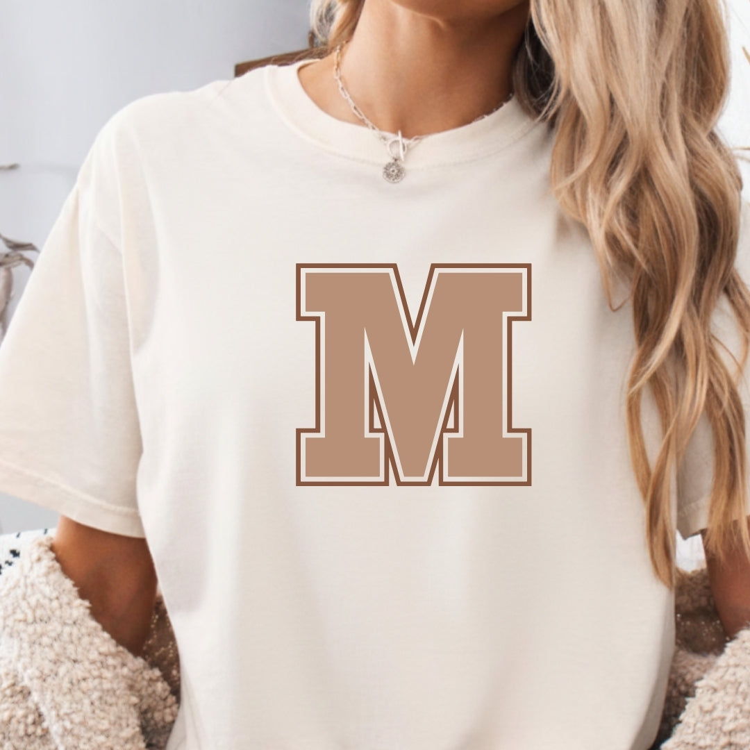 White t-shirt with a brown 'M' on a person wearing a white sweater.
