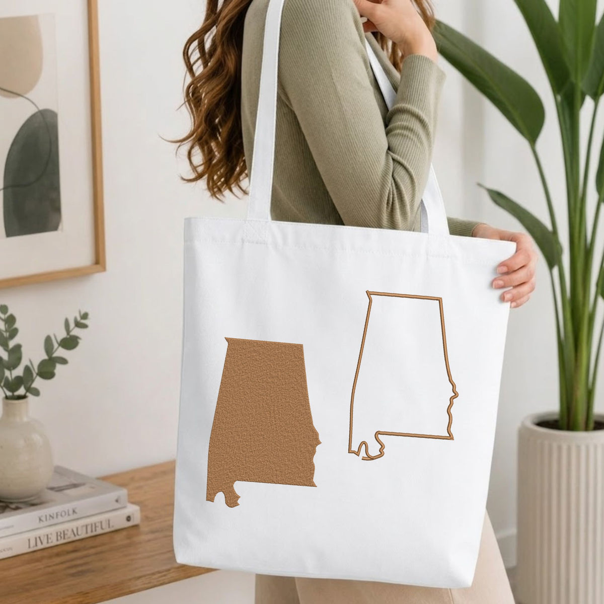 Woman carrying a white natural canvas tote bag featuring an embroidered baby feet in a heart design in a bright, modern interior setting