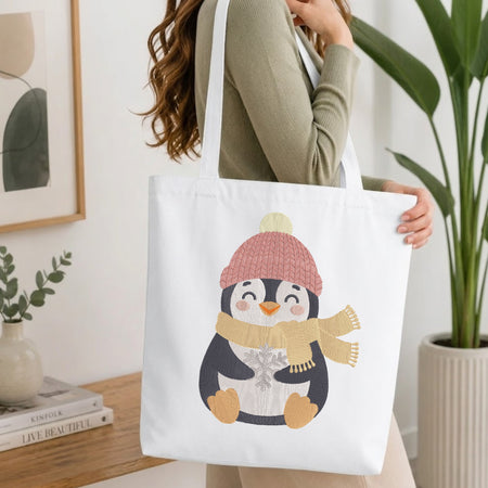 A woman holds a white canvas tote bag featuring a cute penguin illustration wearing a pink knit hat and a beige scarf. The scene is set in a bright, modern room with a wooden table, books, and a potted plant.
