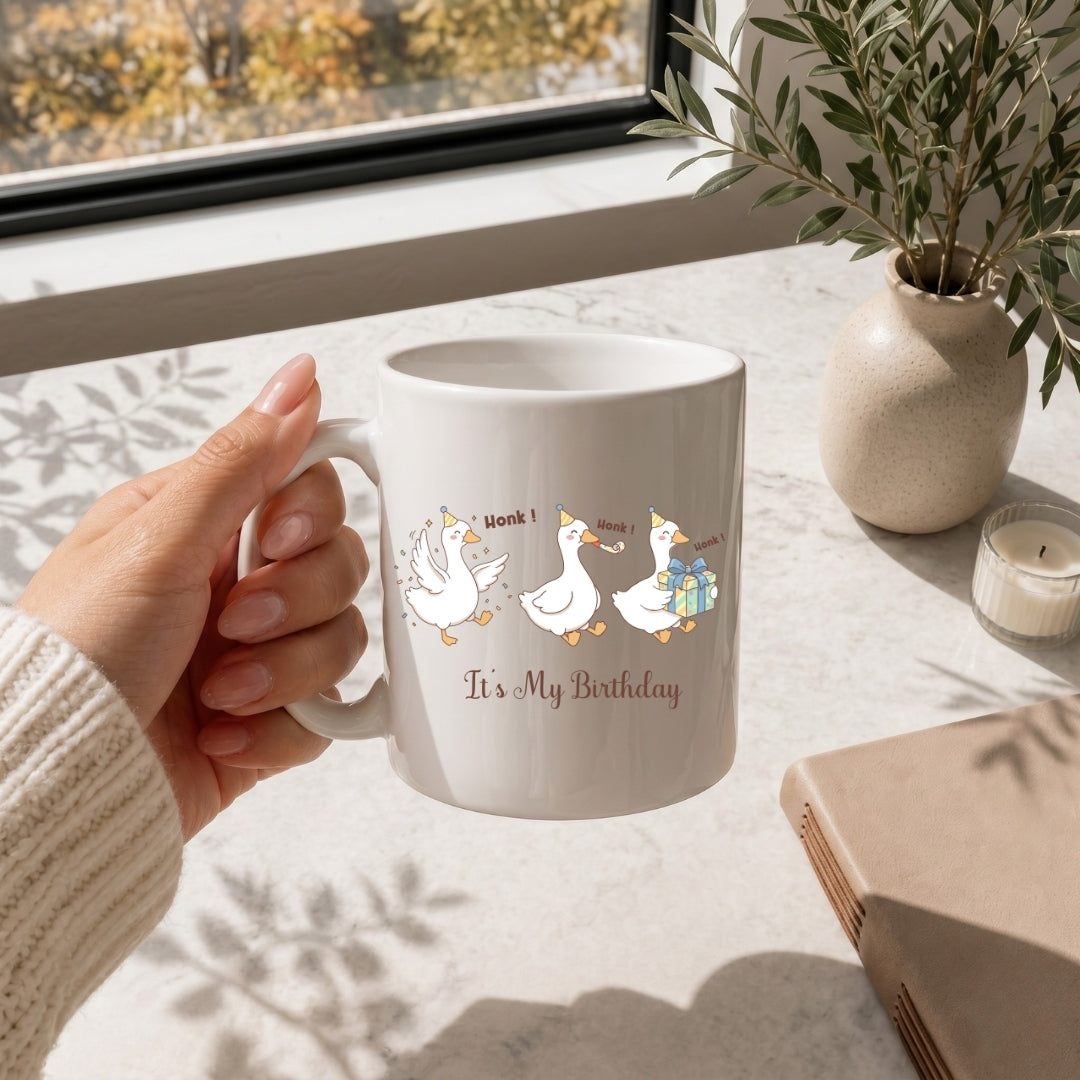 Hand holding a white ceramic coffee mug printed with the cute and funny "It's My Birthday" party geese illustration.