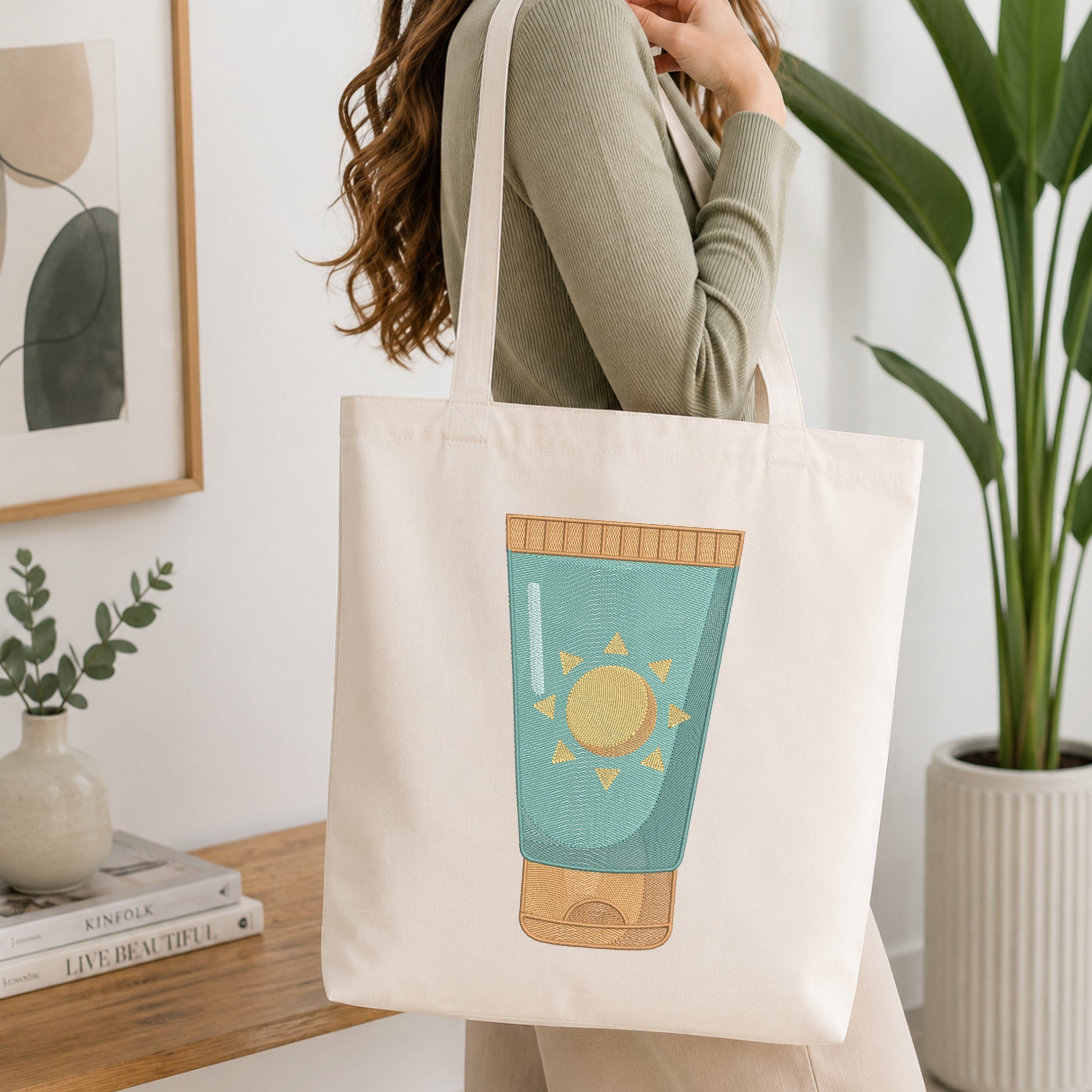 A woman wearing a sage green ribbed long-sleeve top carries a cream canvas tote bag. The bag is off-white with a large graphic illustration of a teal sunscreen tube with a yellow sun on the front.