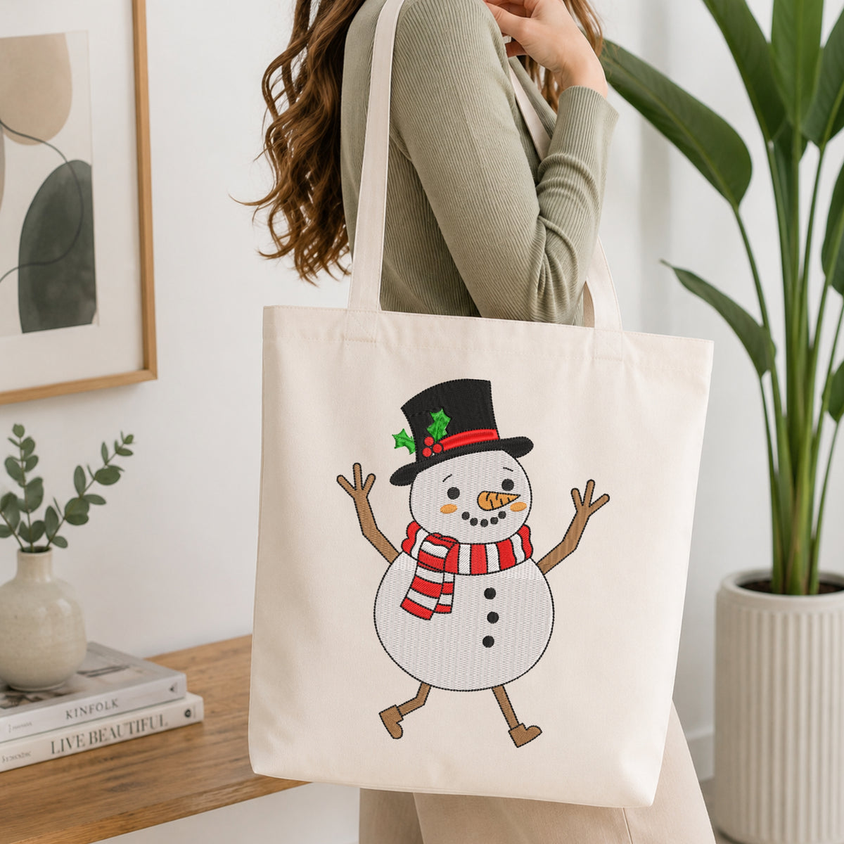 A woman in a ribbed sage green top carries a cream canvas tote bag featuring a cartoon snowman with a black top hat, red and green striped scarf, and orange carrot nose. The scene is set in a bright, modern room with a wooden table, books, and a potted plant.