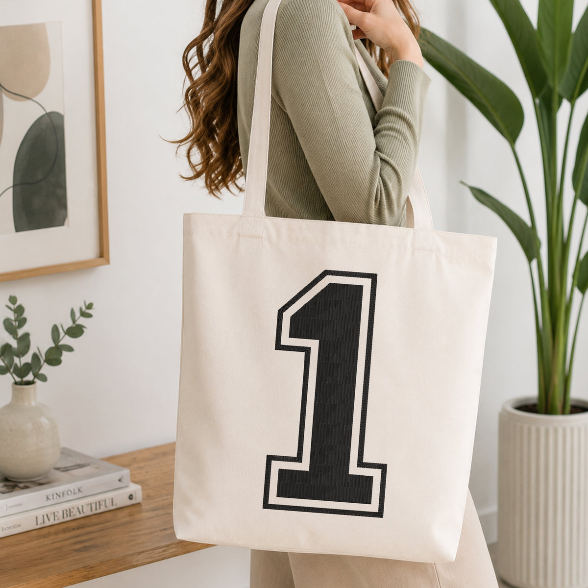 A woman wearing a sage green ribbed long-sleeve top carries a cream canvas tote bag with a large black number 1 outlined in white on the front, set in a bright, modern room with a wooden table, framed art, and a potted plant.