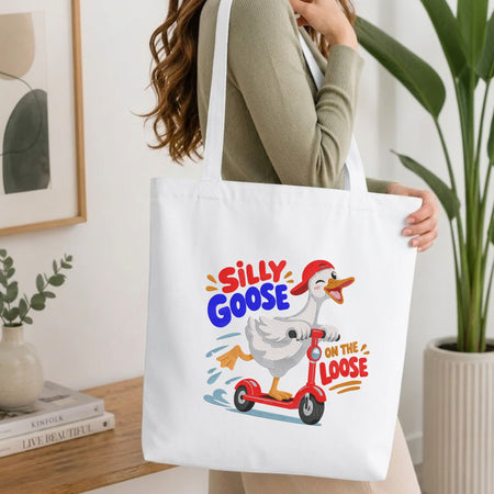  Woman carrying a white natural canvas tote bag featuring a "Silly Goose on the Loose" duck riding a red scooter machine embroidery design in a bright, modern interior setting   
