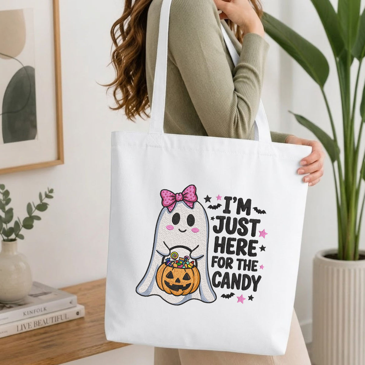 Woman carrying a white natural canvas tote bag featuring an "I'm Just Here for the Candy" cute ghost machine embroidery design in a bright, modern interior setting