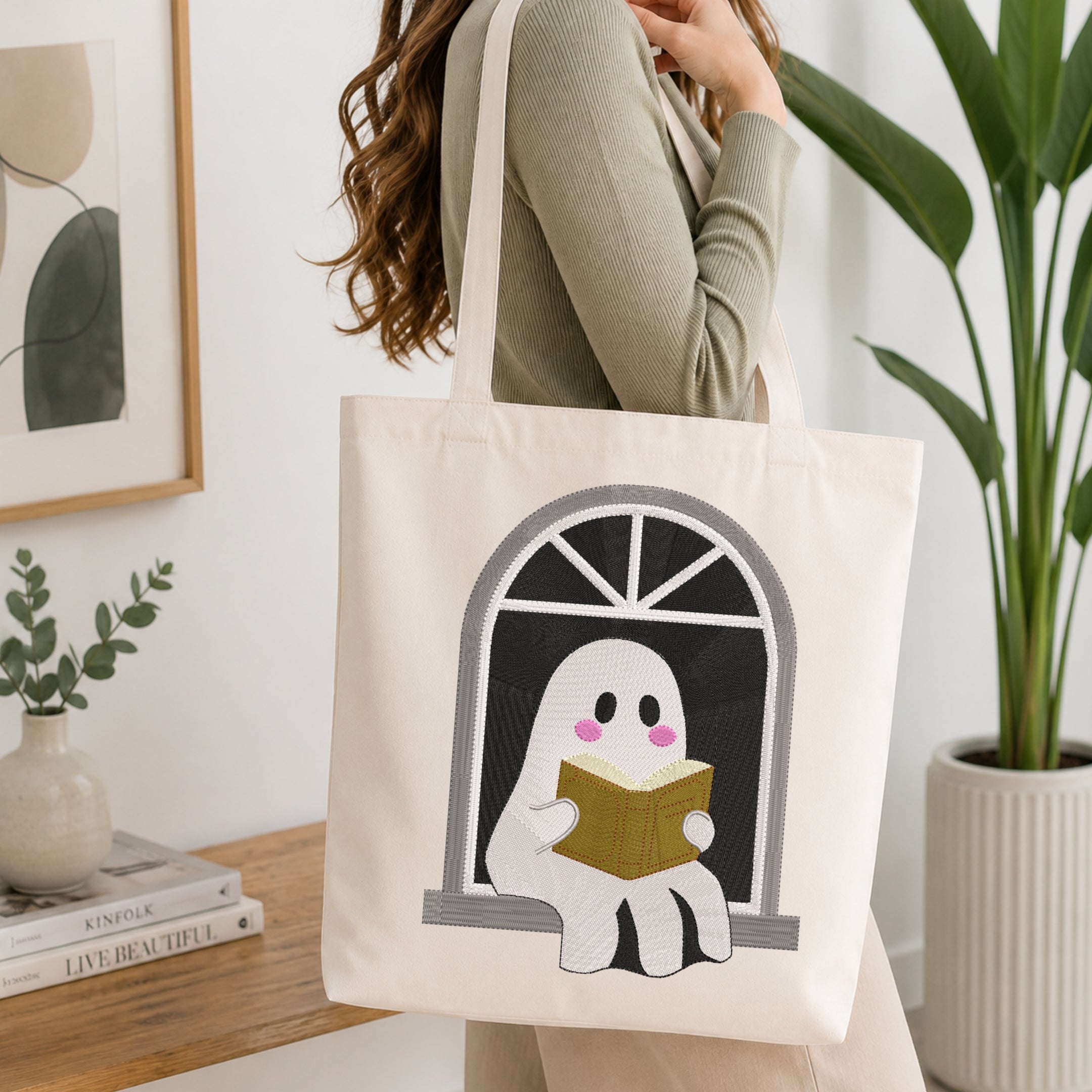 A woman wearing a sage green ribbed sweater carries a cream canvas tote bag with a graphic of a cute white ghost reading a yellow book in a window. The scene is set in a bright, modern room with a wooden table, books, a small plant, and a large green potted plant in the background.