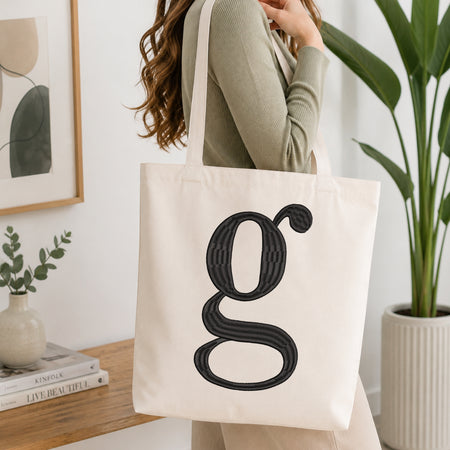 A woman wearing a sage green ribbed long-sleeve top carries a cream canvas tote bag with a large black textured letter 'g' printed on the front, set in a bright, modern room with a wooden console table, framed art, and a potted plant.