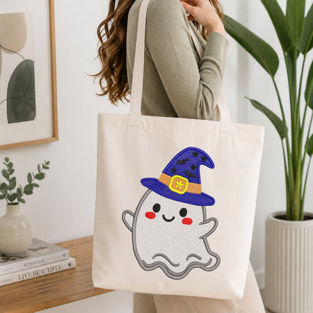 A person wearing a sage green ribbed long-sleeve top carries a cream canvas tote bag. The bag features a cute embroidered-style white ghost wearing a purple wizard hat with a yellow band and black bat details.
