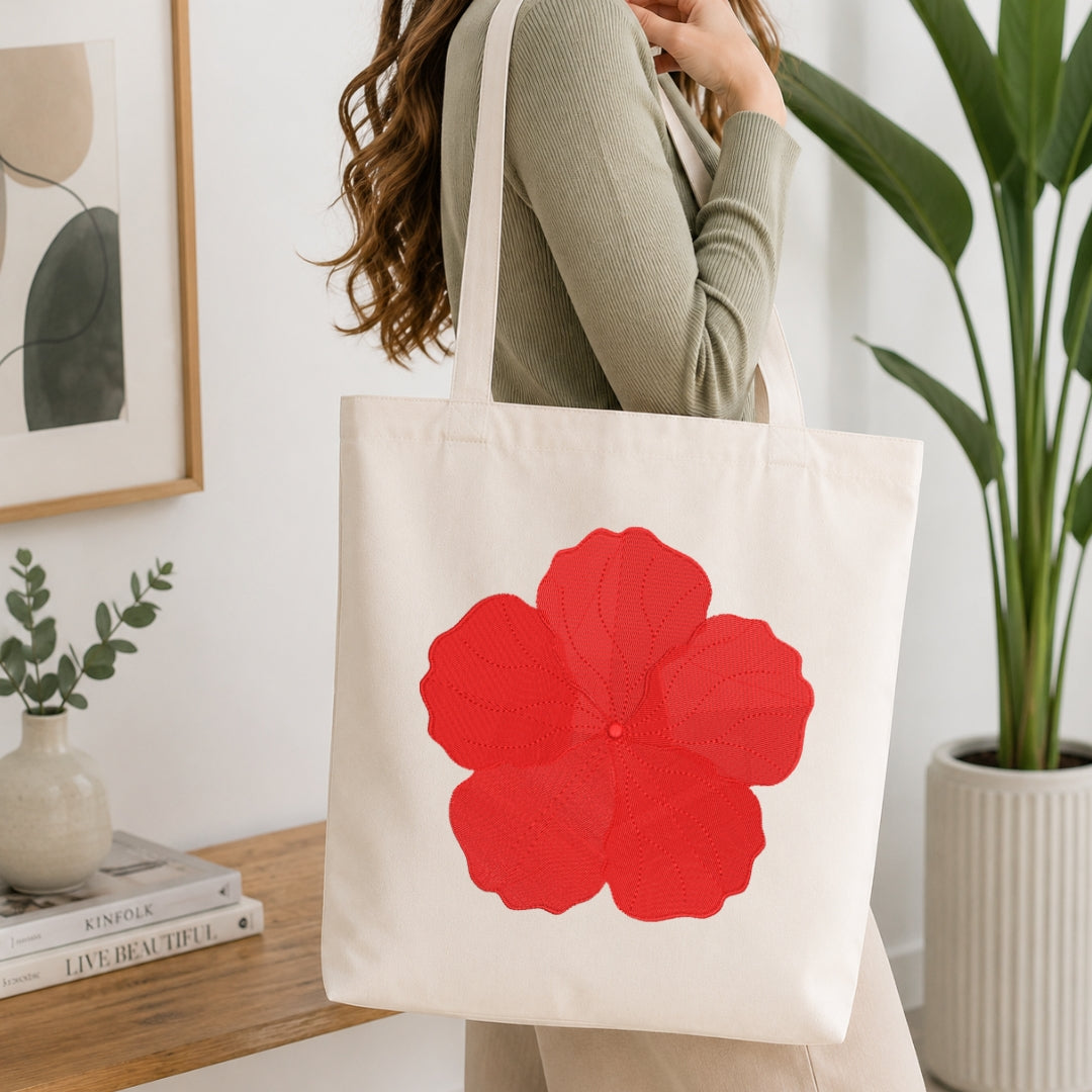 A woman wearing a sage green ribbed sweater carries a cream canvas tote bag with a large red flower graphic on the front, set in a bright, modern room with a wooden table, books, and a potted plant.