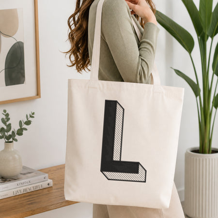 A woman wearing a sage green ribbed long-sleeve top carries a cream canvas tote bag with a large black block letter 'L' printed on the front, set in a bright, modern room with a wooden table, framed art, and a potted plant.