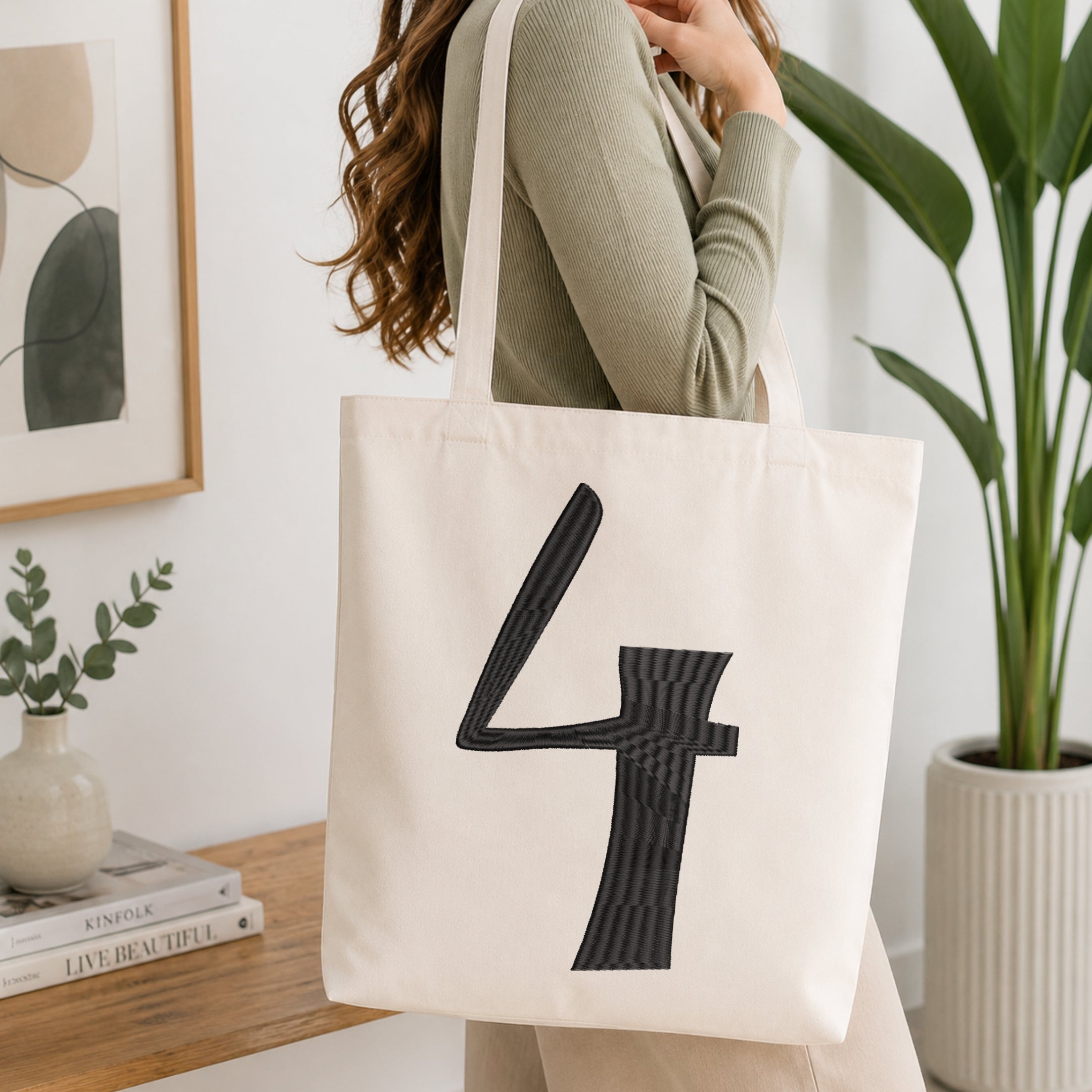 A woman wearing a sage green ribbed top carries a cream canvas tote bag with a large black textured number 4 printed on the front, set in a bright, modern room with a wooden table, framed art, and a potted plant.