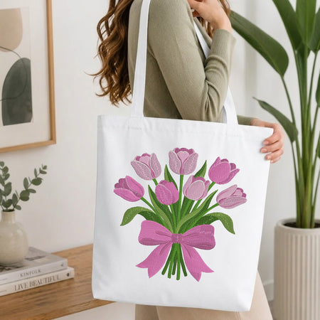 A woman holds a white canvas tote bag featuring a graphic of pink tulips tied with a pink bow and green stems. The scene is set in a bright, modern room with a wooden table, books, and a potted plant in the background.