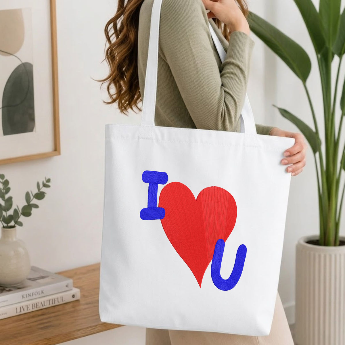 A woman wearing a sage green ribbed long-sleeve top carries a white canvas tote bag with a colorful 'I [red heart] U' graphic print. The setting is a bright, modern room with a wooden table, books, a framed print, and a potted plant.