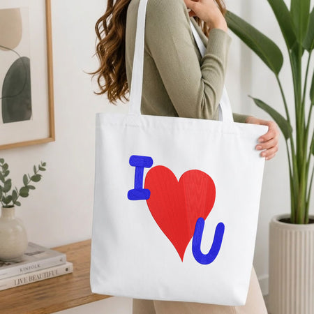 A woman wearing a sage green ribbed long-sleeve top carries a white canvas tote bag with a colorful 'I [red heart] U' graphic print. The setting is a bright, modern room with a wooden table, books, a framed print, and a potted plant.