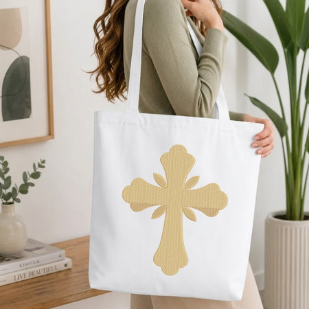 A woman wearing a sage green ribbed long-sleeve top carries a white canvas tote bag with a large textured gold cross design on the front, set in a bright, modern room with a wooden table, books, and a potted plant.
