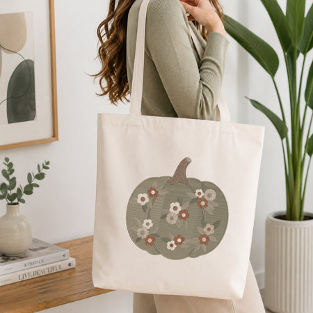 A woman wearing a sage green ribbed long-sleeve top carries a cream canvas tote bag with a large embroidered pumpkin design featuring white and pink flowers. The scene is set in a bright, modern room with a wooden table, books, a framed print, and a potted plant.