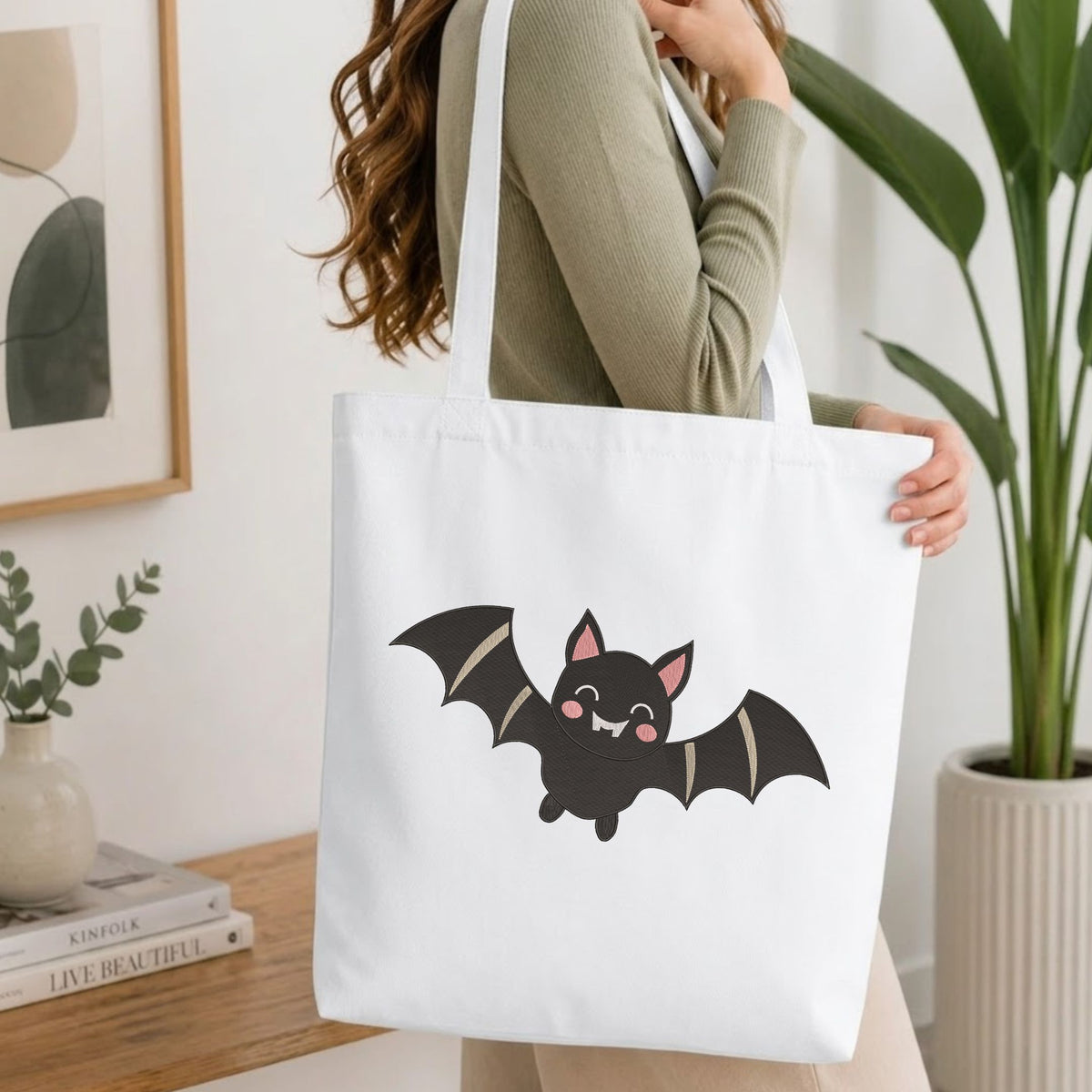 Woman carrying a white natural canvas tote bag featuring an embroidered cute Halloween bat design in a bright, modern interior setting.