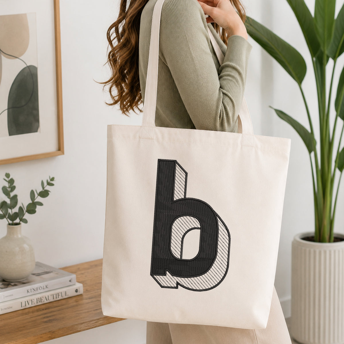 A woman wearing a sage green ribbed long-sleeve top carries a cream canvas tote bag with a large black stylized letter 'b' printed on the front, set in a bright, modern room with a wooden console table and a potted plant.