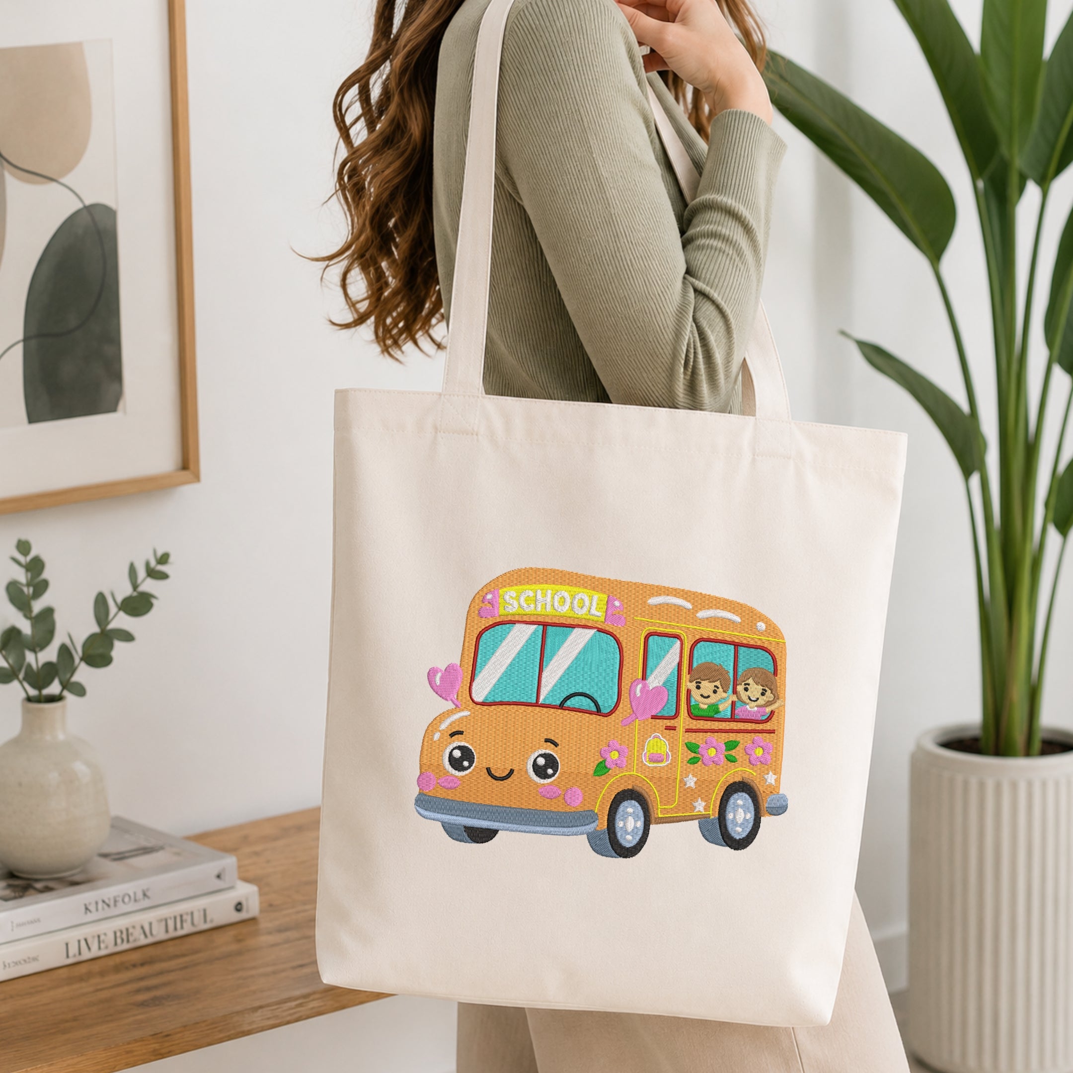 A woman wearing a sage green ribbed sweater carries a cream canvas tote bag with a colorful cartoon school bus graphic. The tote bag features a smiling orange bus with pink hearts, blue windows, and a green 'SCHOOL' sign.