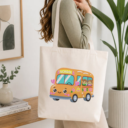 A woman wearing a sage green ribbed sweater carries a cream canvas tote bag with a colorful cartoon school bus graphic. The tote bag features a smiling orange bus with pink hearts, blue windows, and a green 'SCHOOL' sign.