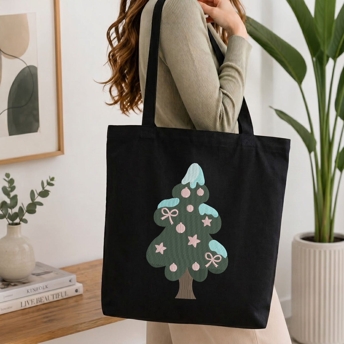 A woman wearing a beige ribbed sweater carries a black canvas tote bag with a stylized green Christmas tree graphic decorated with pink ornaments and light blue accents, set against a bright, modern indoor background.