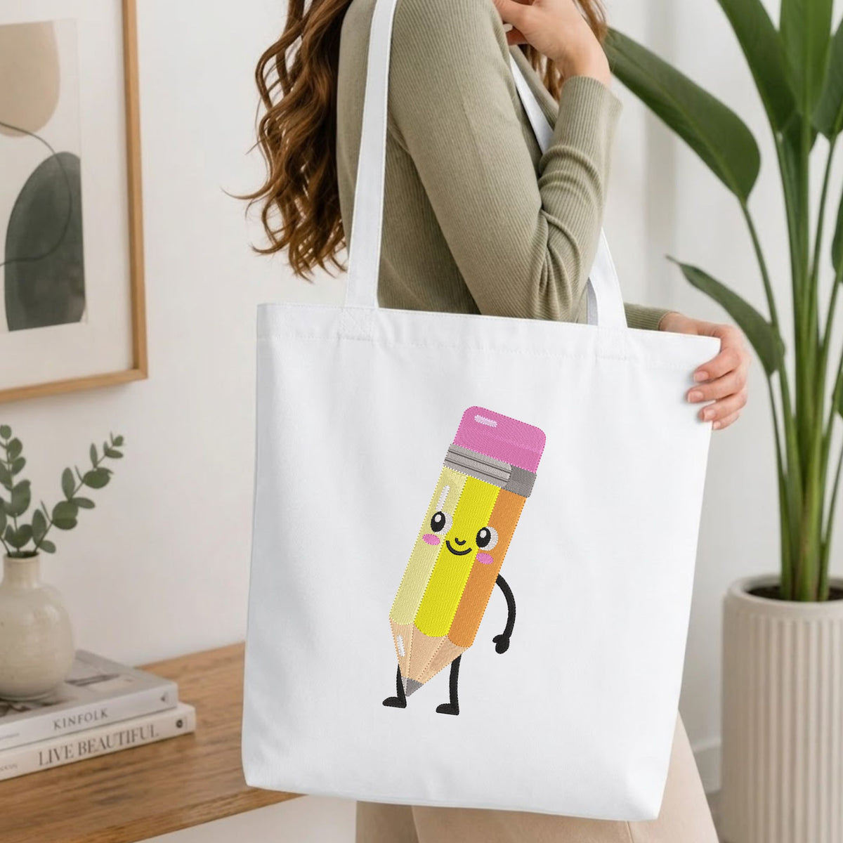 A woman wearing a sage green ribbed long-sleeve top carries a white canvas tote bag. The bag features a cute cartoon graphic of a smiling yellow pencil with a pink eraser cap and black limbs.