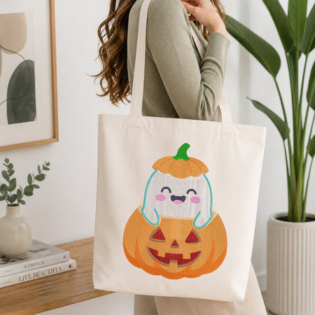 A person wearing a sage green ribbed long-sleeve top carries a cream canvas tote bag with a cute cartoon ghost jack-o'-lantern graphic. The tote bag is shown in a bright, modern room setting with a wooden table, books, a framed print, and a potted plant.