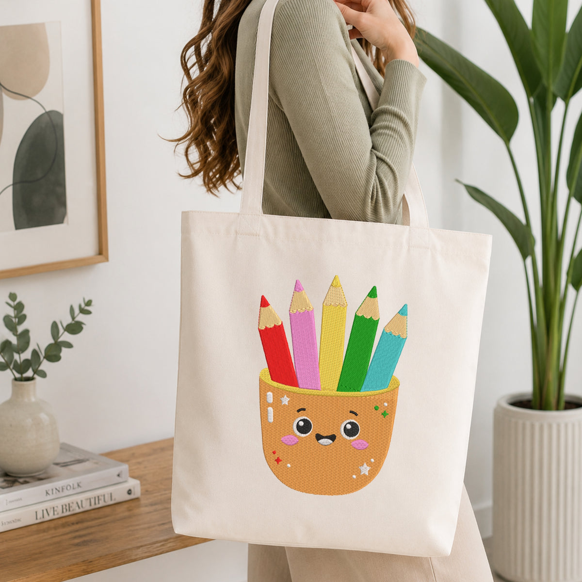 A woman wearing a sage green ribbed long-sleeve top carries a cream canvas tote bag. The bag features a cute graphic of a smiling orange pencil cup filled with colorful pencils (red, pink, yellow, green, and blue).