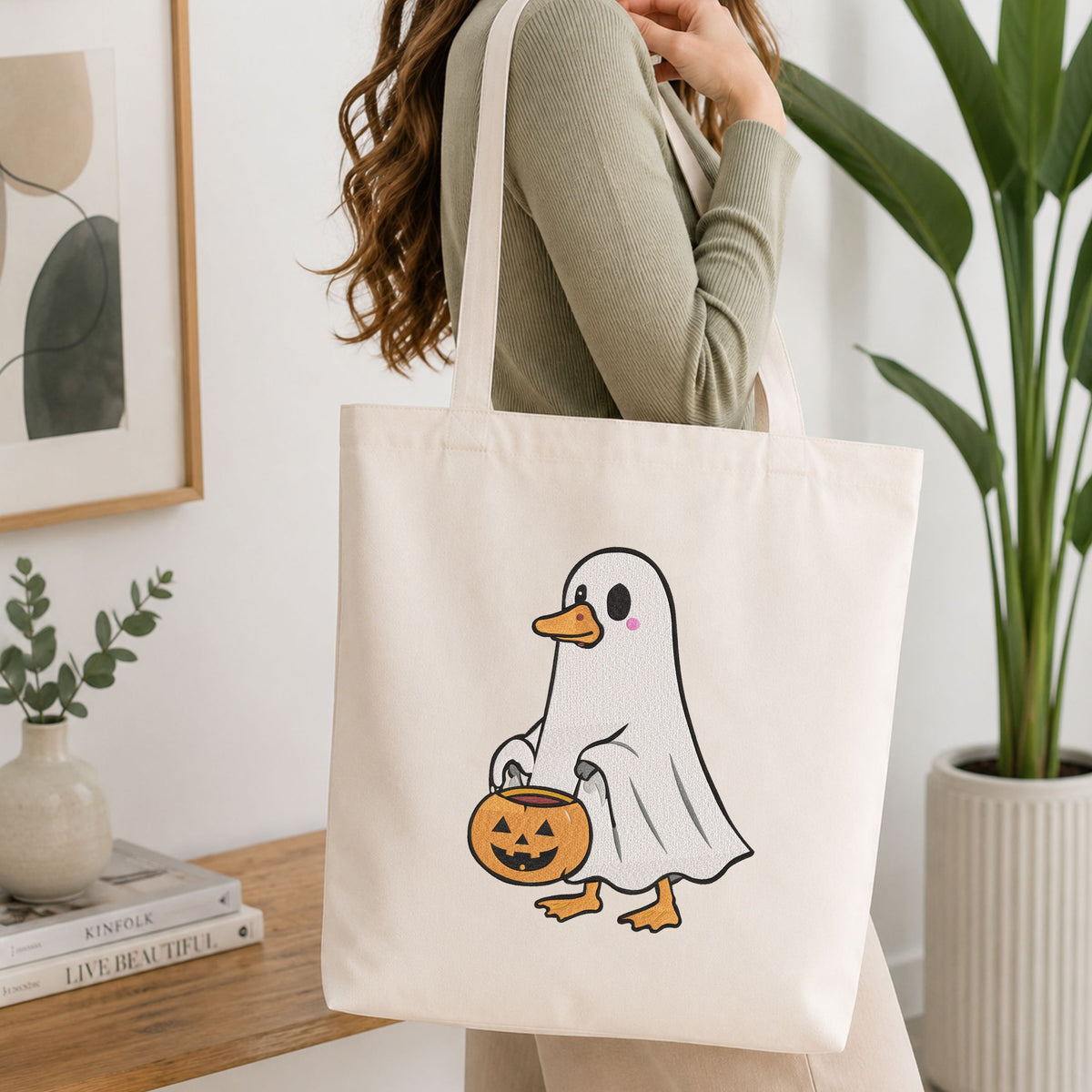 Woman carrying a white natural canvas tote bag featuring a cute duck in a ghost costume holding a trick-or-treat bucket machine embroidery design in a bright, modern interior setting