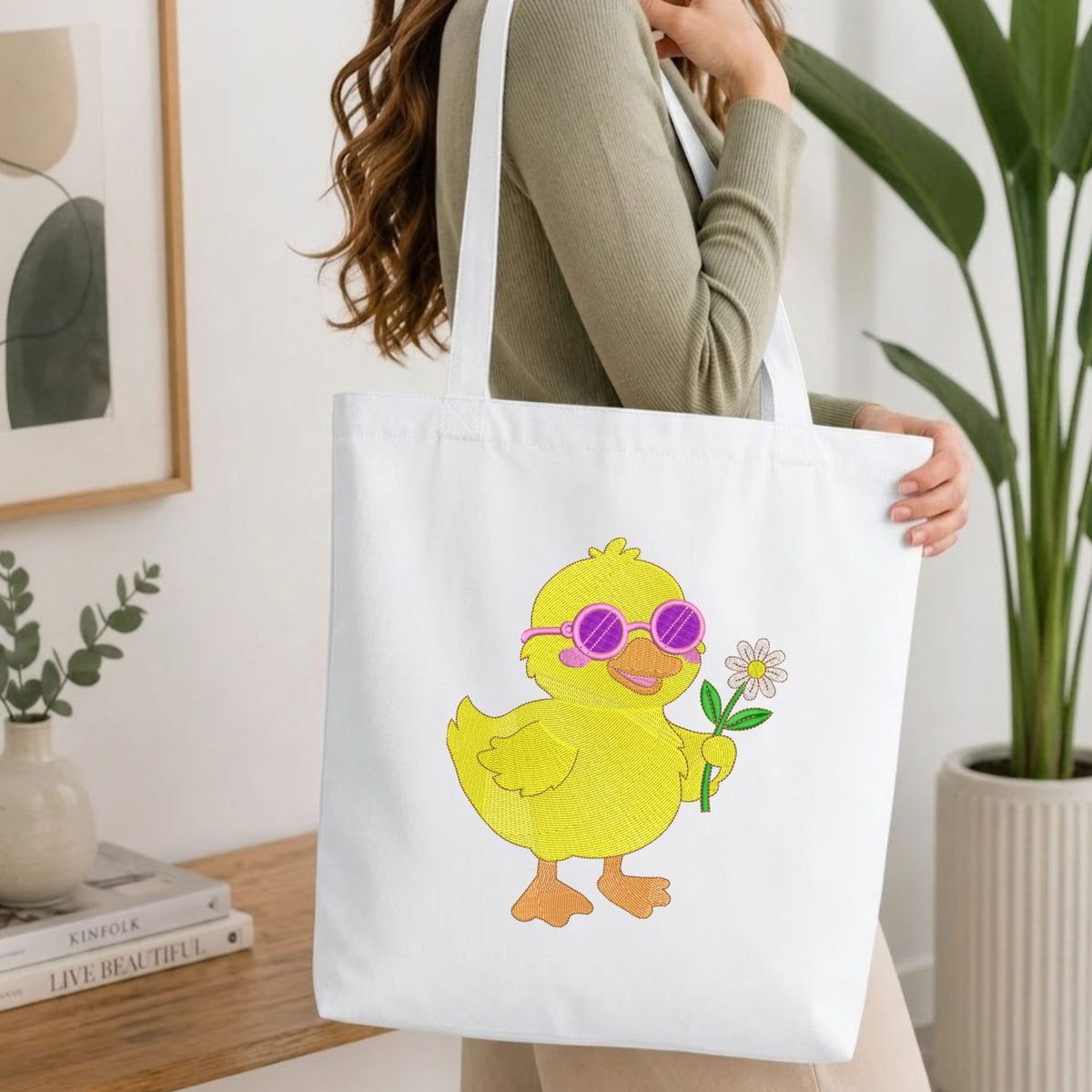 Folded white cotton t-shirt mockup featuring a cute yellow duck with sunglasses machine embroidery design, styled with cotton stems and a wooden cup.