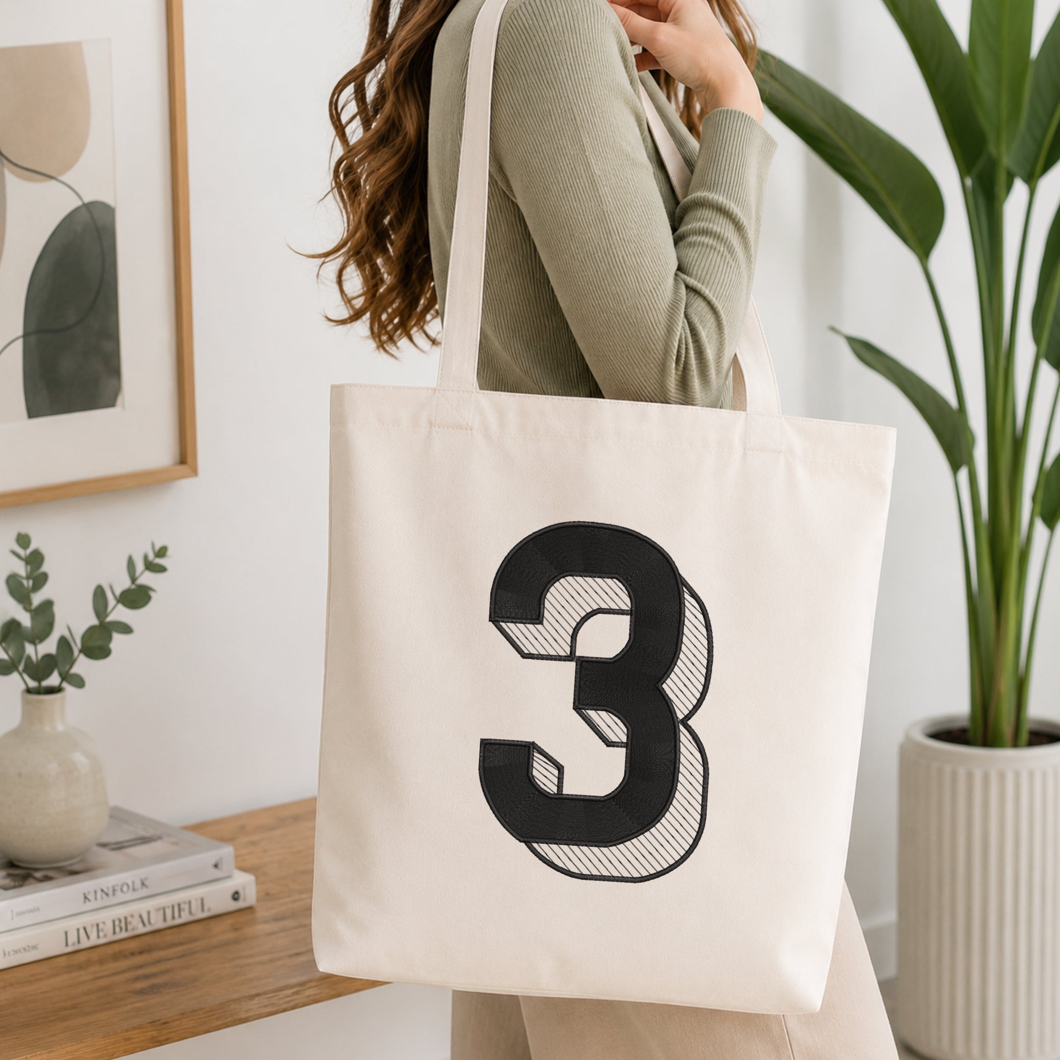 A woman wearing a sage green ribbed long-sleeve top carries a cream canvas tote bag with a large black number 3 printed on the front, set in a bright, modern room with a wooden table, framed art, and a potted plant.