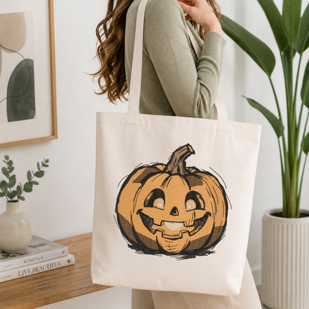A person wearing a sage green ribbed long-sleeve top carries a cream canvas tote bag with a hand-drawn orange jack-o'-lantern pumpkin graphic on the front, set in a bright, modern room with a wooden table, books, and a potted plant.