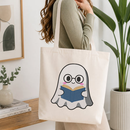 A woman wearing a sage green ribbed long-sleeve top carries a cream canvas tote bag with a cute cartoon ghost graphic reading a blue book, set in a bright, modern room with a wooden table, framed art, and a potted plant.