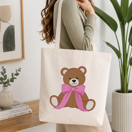 A person wearing a sage green ribbed sweater carries a cream canvas tote bag featuring a brown teddy bear with a pink bow on the front, set against a bright, modern interior background with a wooden table, framed art, and a potted plant.
